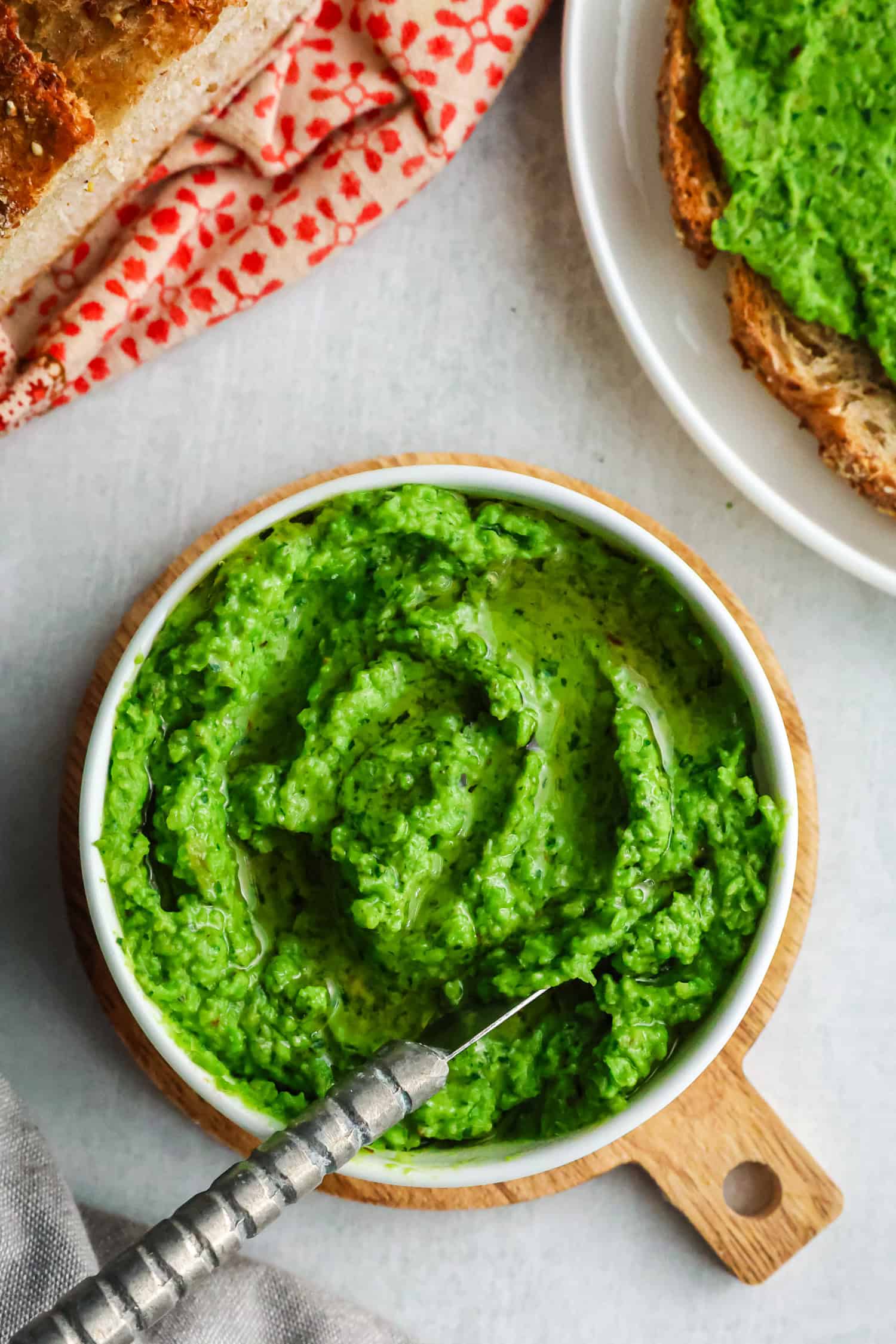 A white bowl filled with chunky green pea spread sitting on a wooden board next to a piece of toast.