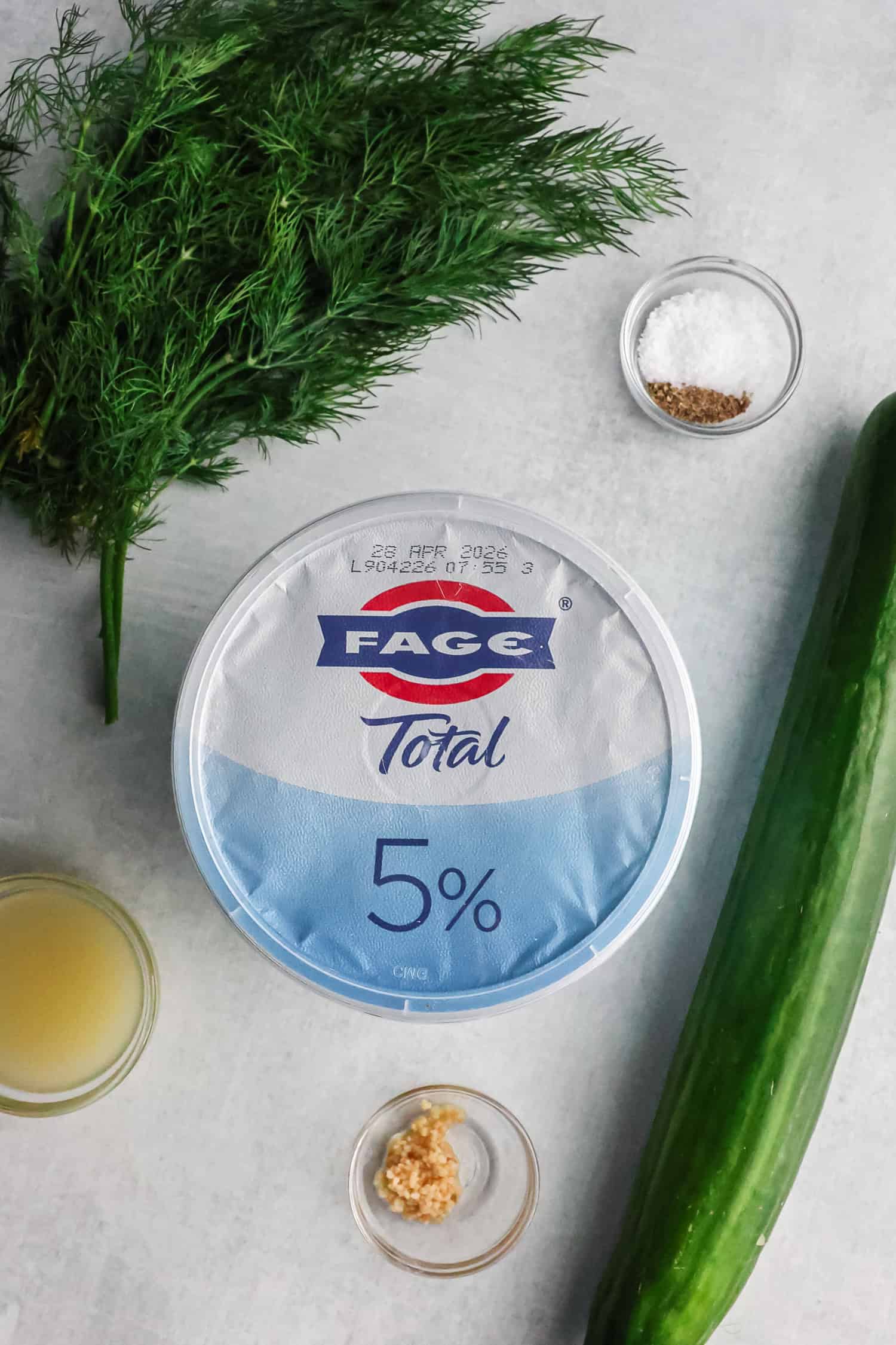 A container of Fage Greek yogurt surrounded by fresh dill, a long cucumber, lemon juice, and minced garlic.