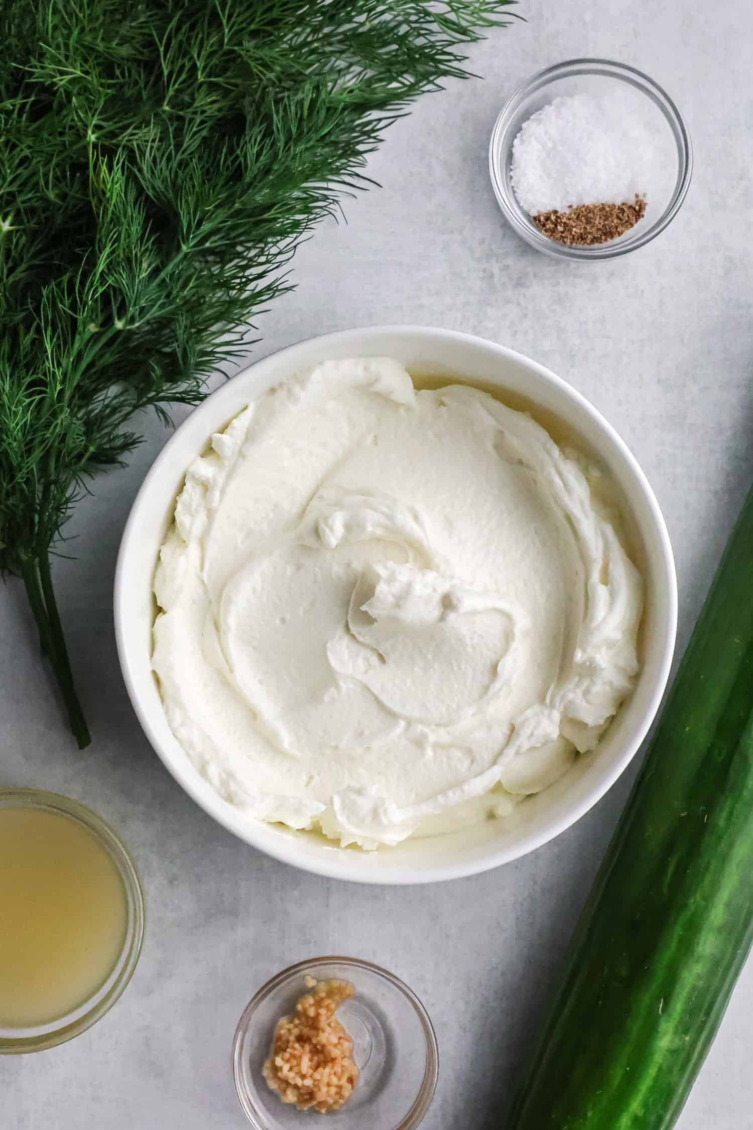A container of Fage Greek yogurt surrounded by fresh dill, a long cucumber, lemon juice, and minced garlic.