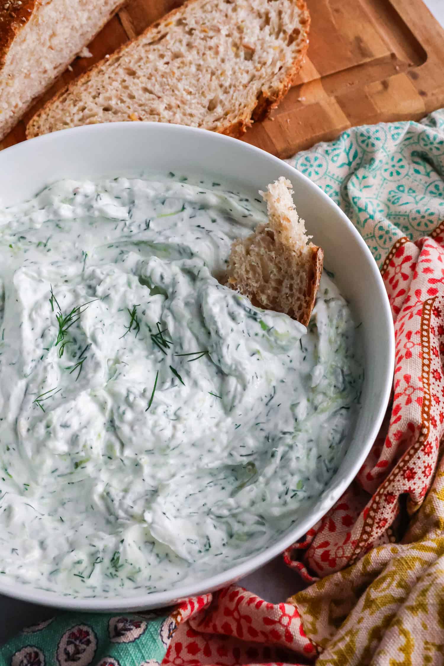 A piece of crusty bread is dipped into a white bowl of creamy yogurt cucumber sauce with fresh dill.