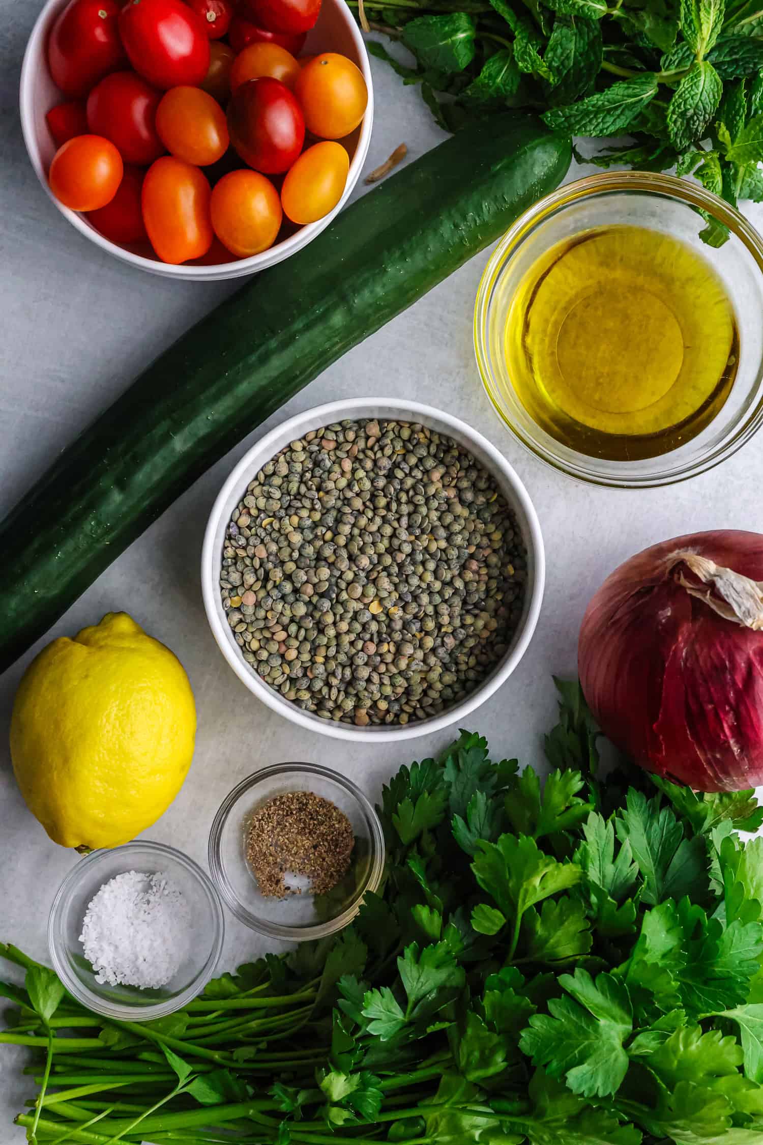 A bowl of dry green lentils sits on a table with fresh ingredients like tomatoes, cucumber, lemon, and herbs.