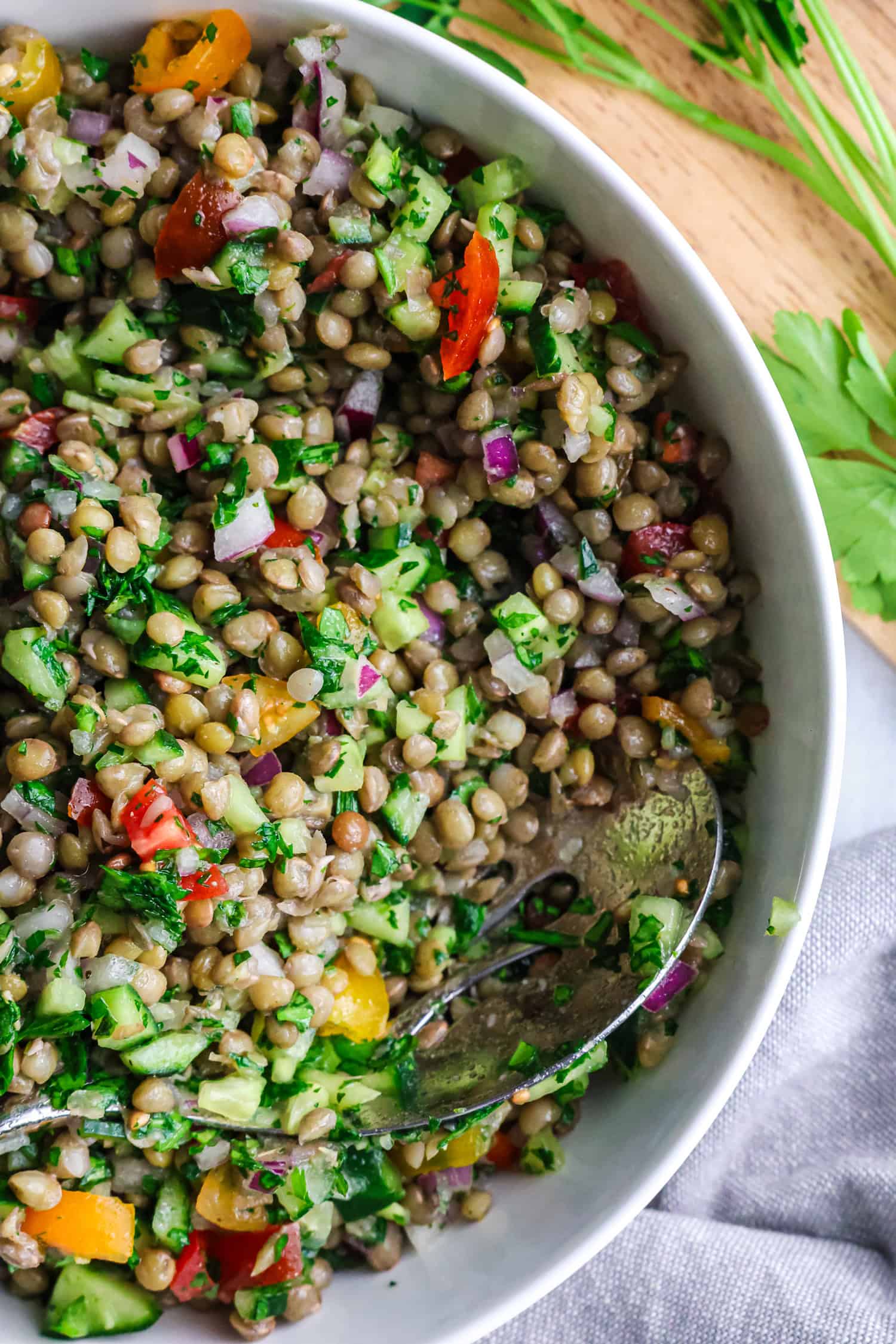 A close-up shot shows a white bowl filled with a fresh salad of lentils, chopped cucumbers, tomatoes, and red onions.