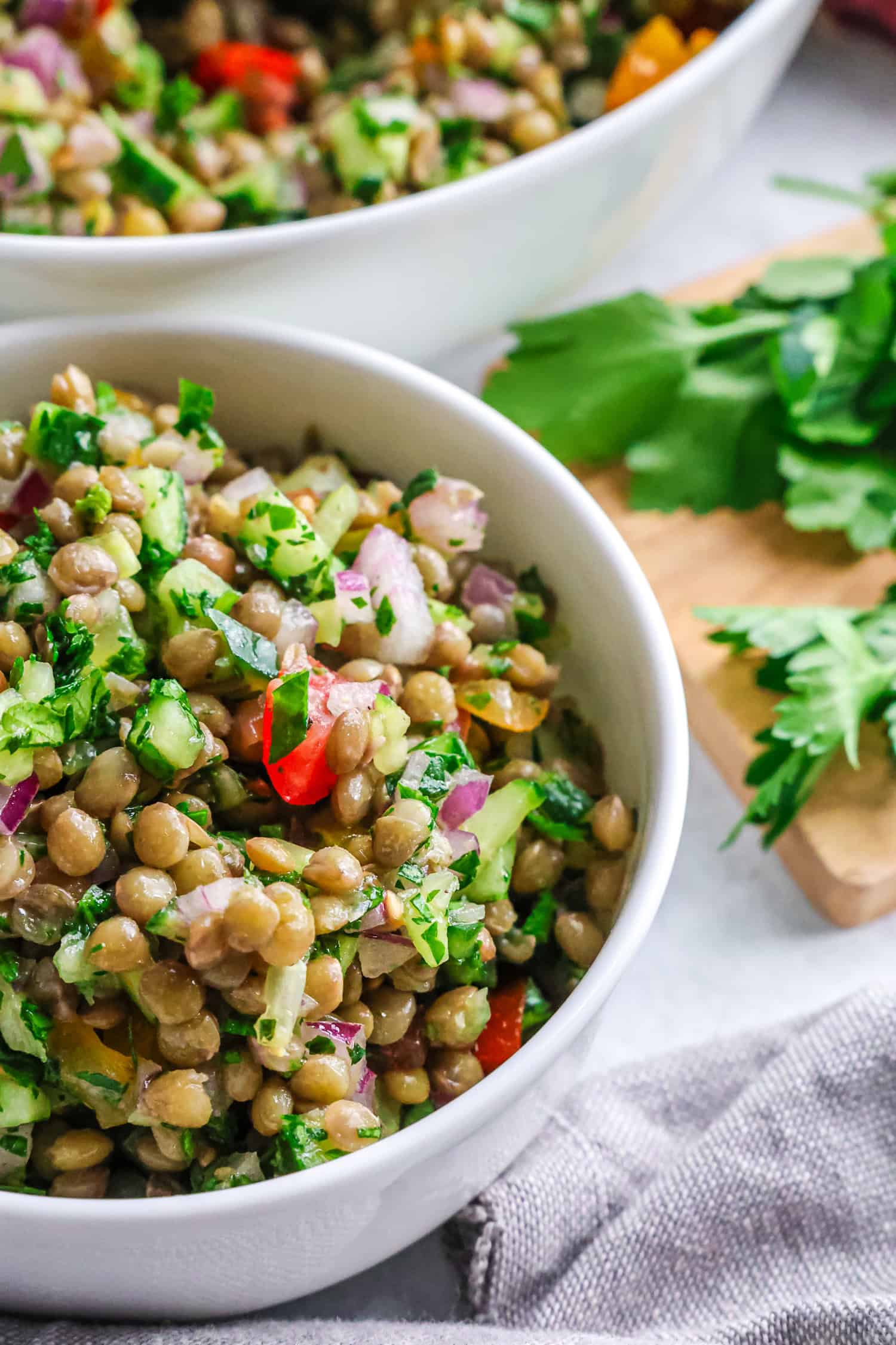 A small white bowl is filled with a fresh lentil and vegetable salad sitting in front of a larger bowl.