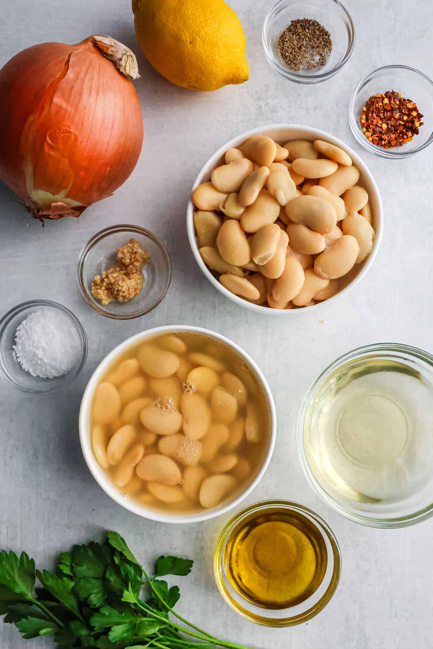 A flat lay of ingredients for a butter bean recipe, including two bowls of canned butter beans (one drained, one in liquid), a whole onion, a lemon, a bunch of parsley, garlic, olive oil, and small bowls of salt, pepper, and red pepper flakes.