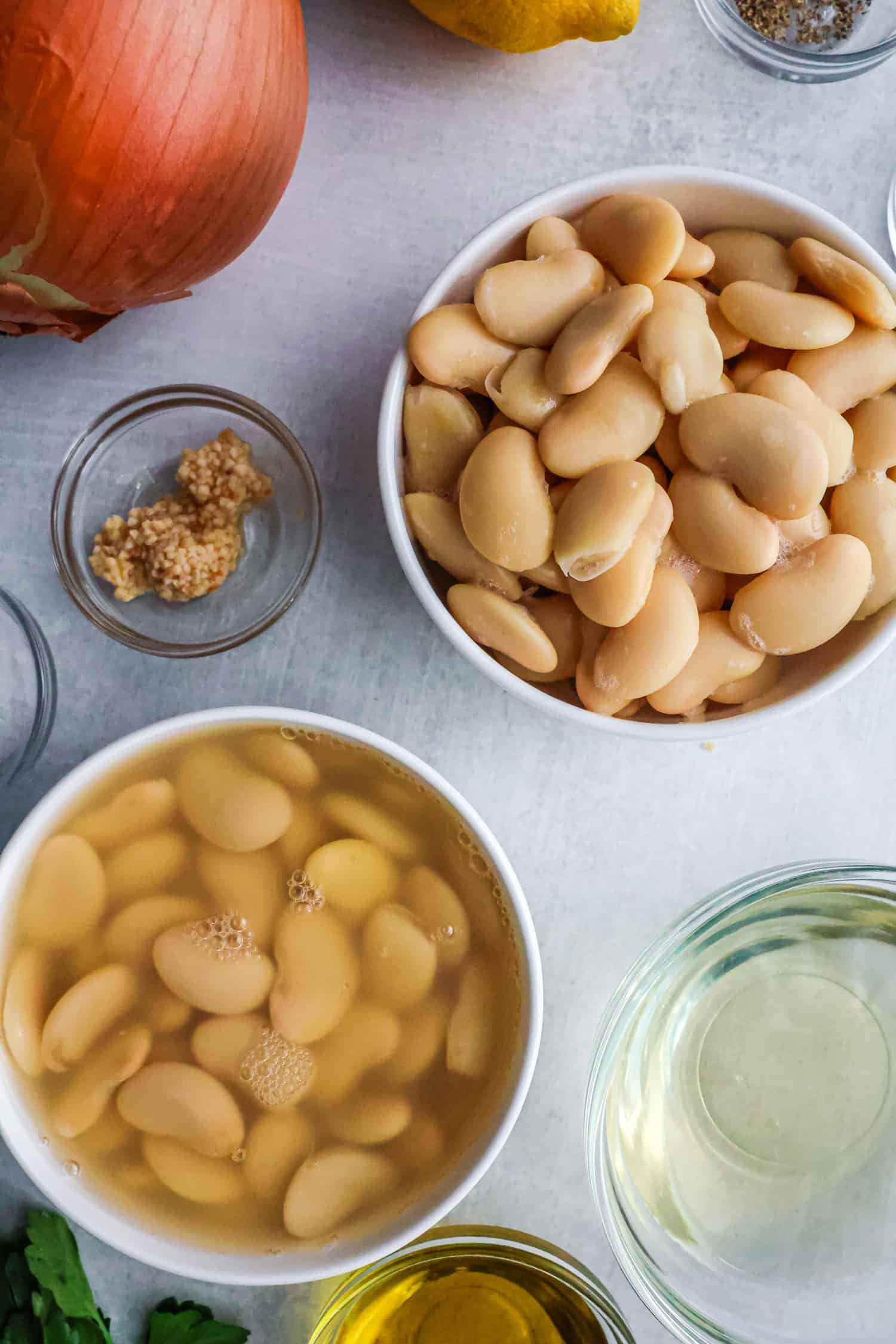Top-down view of two white bowls filled with canned butter beans, surrounded by a whole onion, minced garlic, olive oil, and fresh parsley on a light grey countertop.