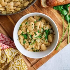 A white bowl filled with cooked butter beans garnished with fresh parsley, sitting on a wooden cutting board next to a piece of crusty bread and a sprig of parsley.