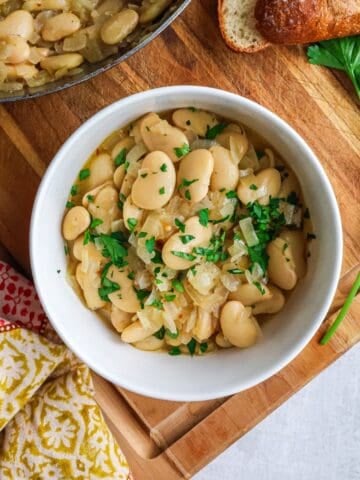A white bowl filled with cooked butter beans garnished with fresh parsley, sitting on a wooden cutting board next to a piece of crusty bread and a sprig of parsley.
