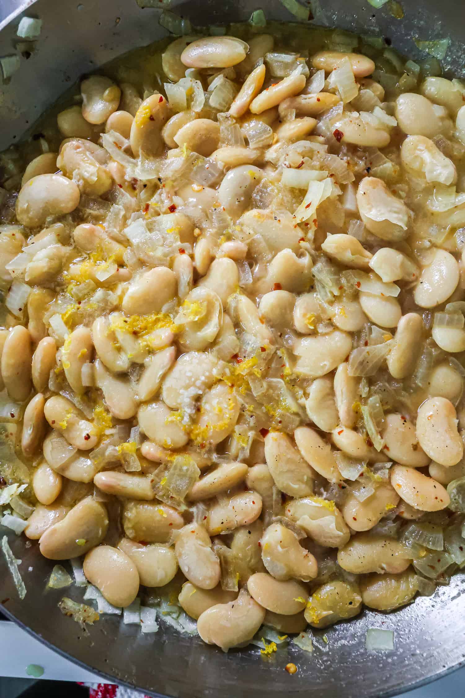 A top-down view of a stainless steel pan filled with cooked butter beans, sautéed onions, red chili flakes, and bright yellow lemon zest in a light, oily sauce.