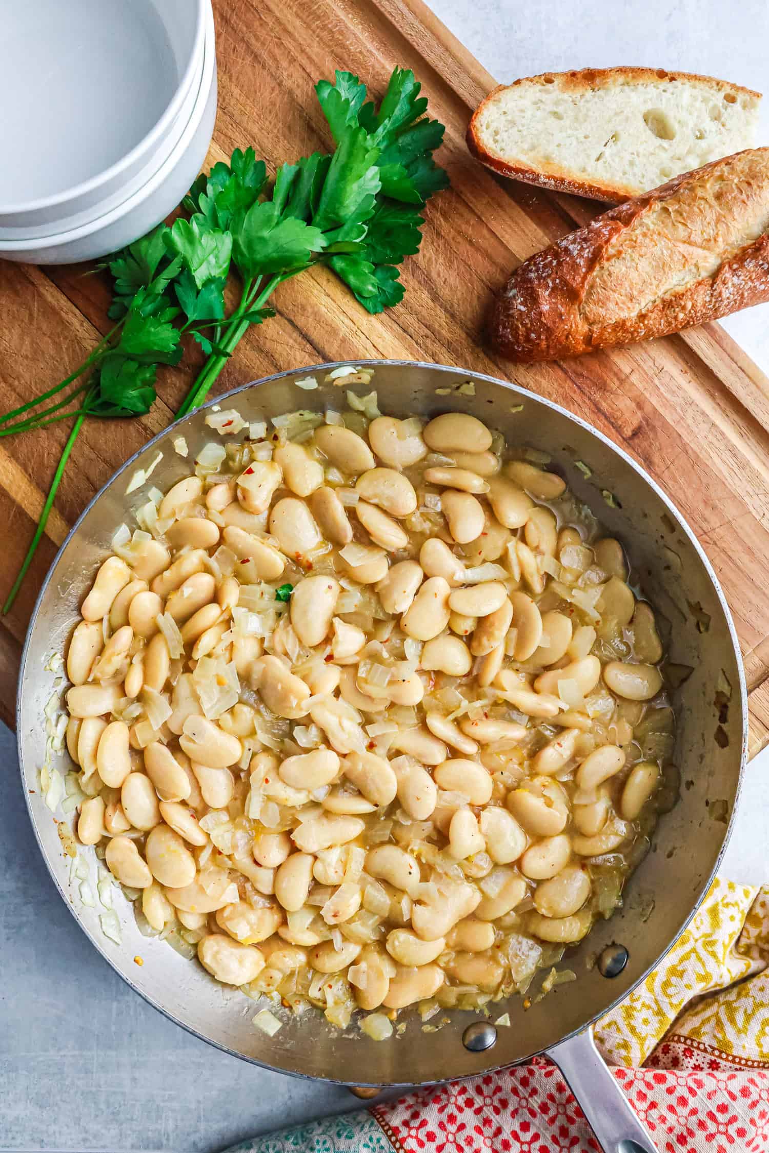 A top-down view of a silver skillet filled with sautéed butter beans and onions, positioned on a light gray surface next to a wooden cutting board with crusty bread, a bunch of fresh parsley, and a stack of white bowls.