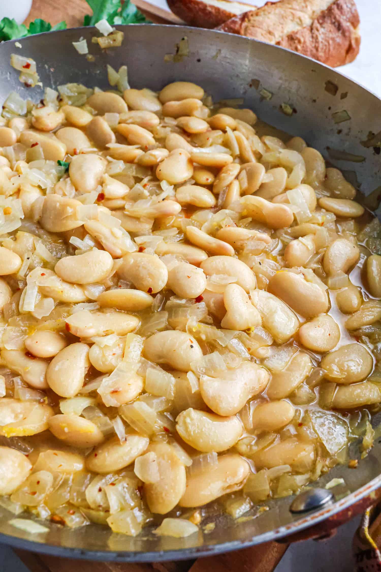 A vertical close-up of a stainless steel skillet filled with large butter beans and sautéed onions in a golden, shimmering sauce, with a crusty baguette loaf visible in the soft-focus background.