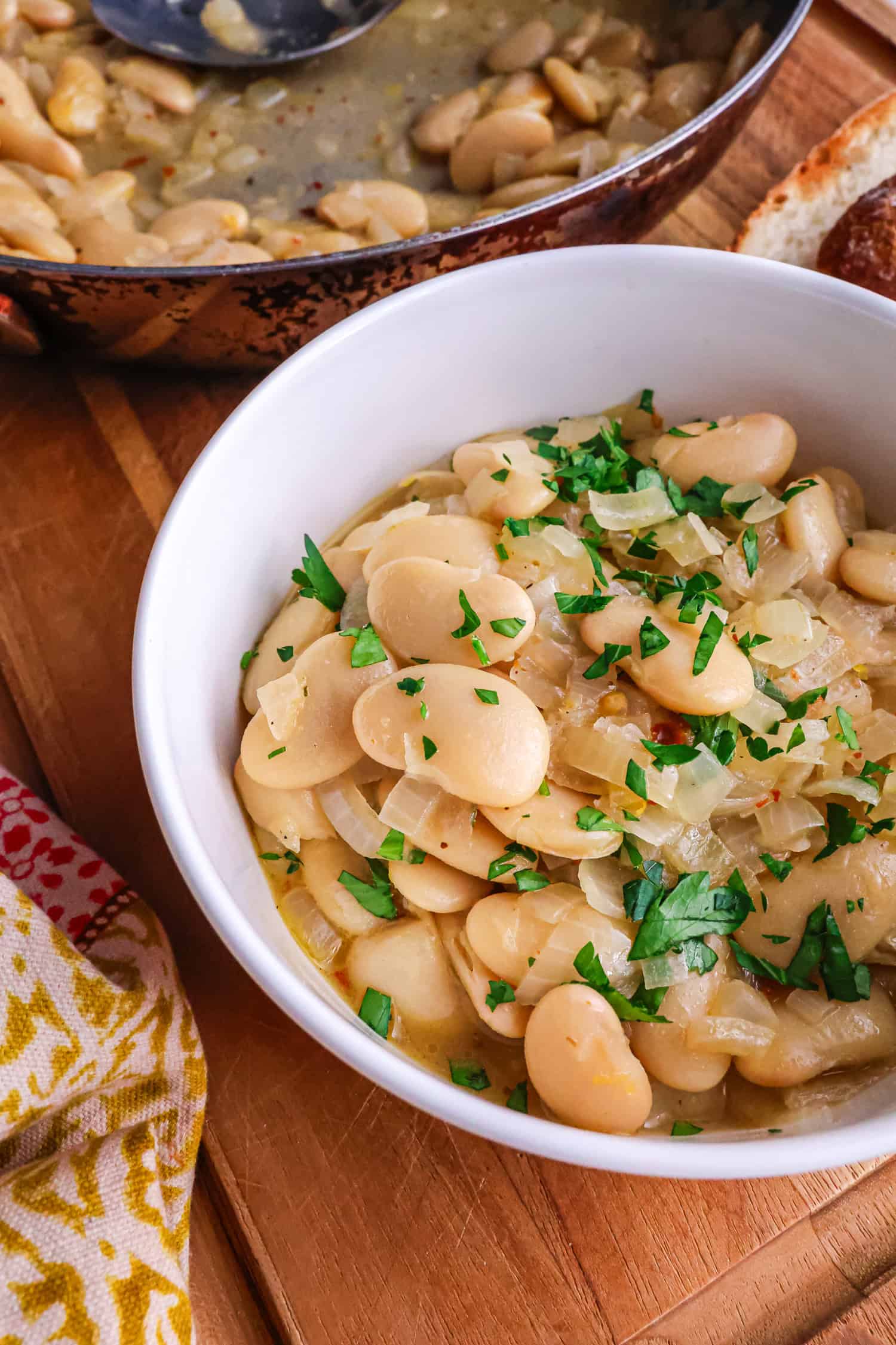 A close-up shot of a white ceramic bowl filled with cooked butter beans and sautéed onions, topped with freshly chopped parsley, with a skillet and crusty bread in the soft-focus background.
