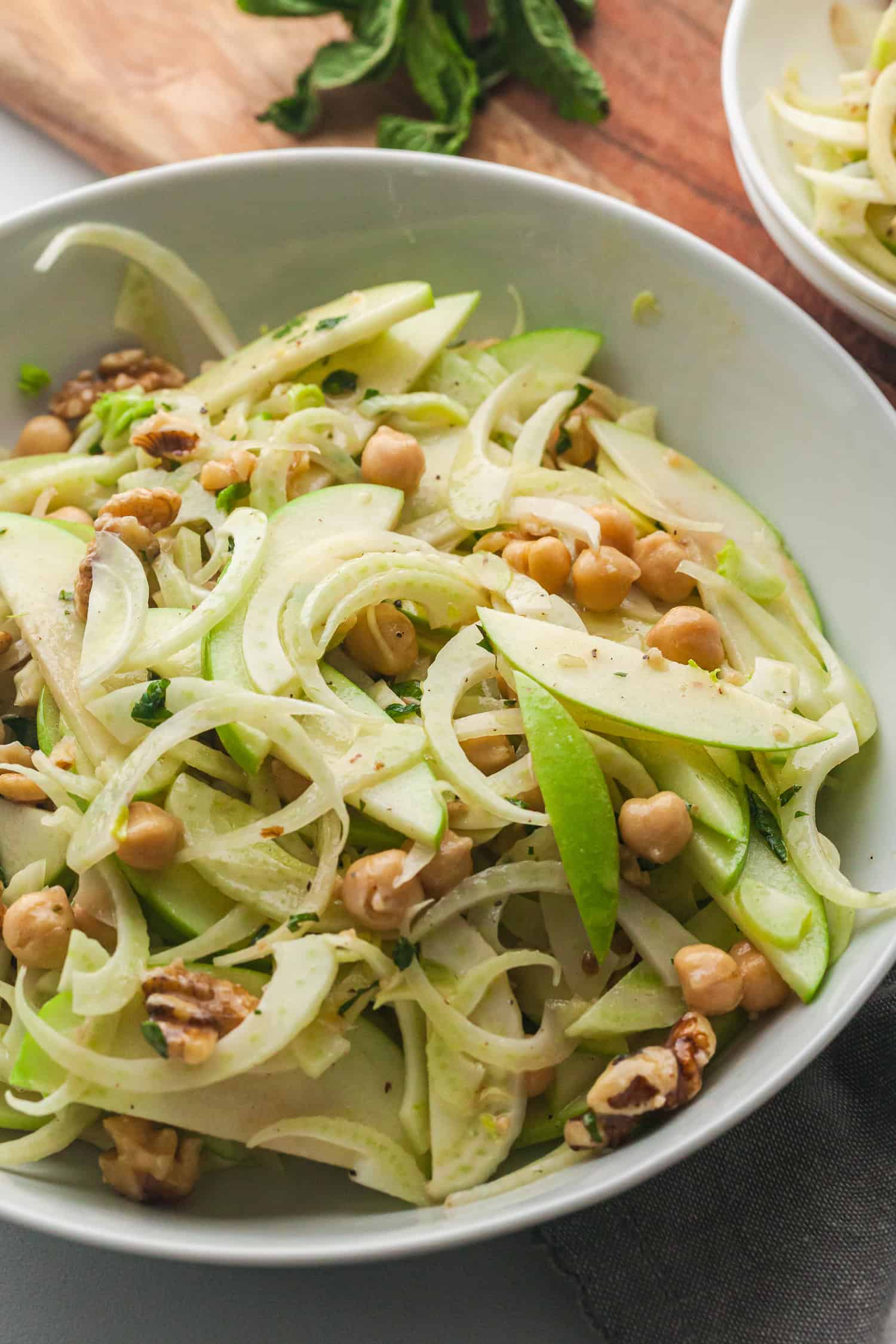 Apple fennel slaw in a white bowl with chickpeas, walnuts, and fresh herbs on a cutting board.
