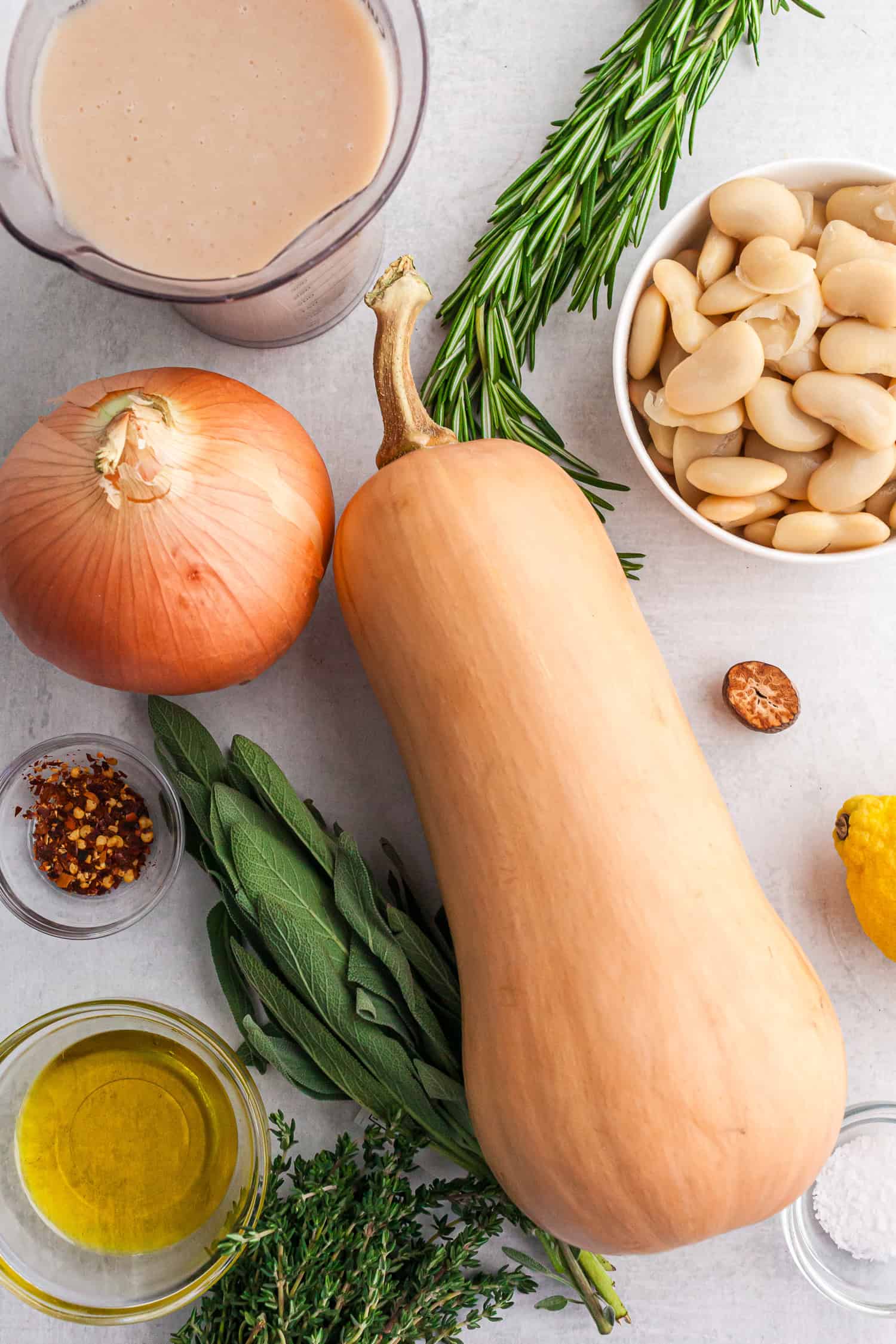 A big butternut squash, a bowl of white beans, fresh rosemary and sage, and olive oil. They are ready to be cooked.