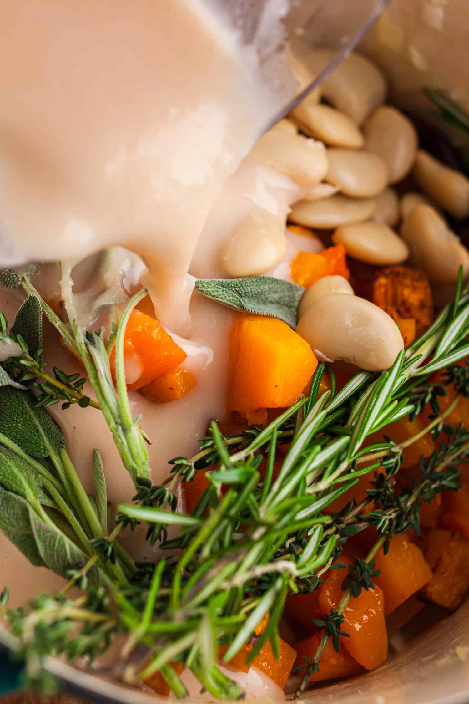 White beans, roasted squash, and fresh herbs in a bowl with creamy broth being poured. The mix looks warm and ready to cook, showing a cozy step in a plant-based recipe.
