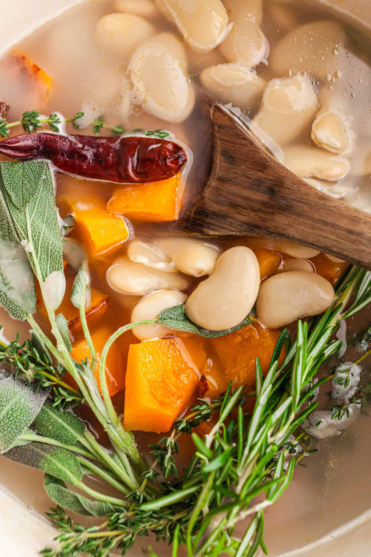 A pot of white beans, orange veggies, herbs, and a red chili cooking in clear broth. A wooden spoon stirs the mix, showing a warm and healthy step in a homemade soup.