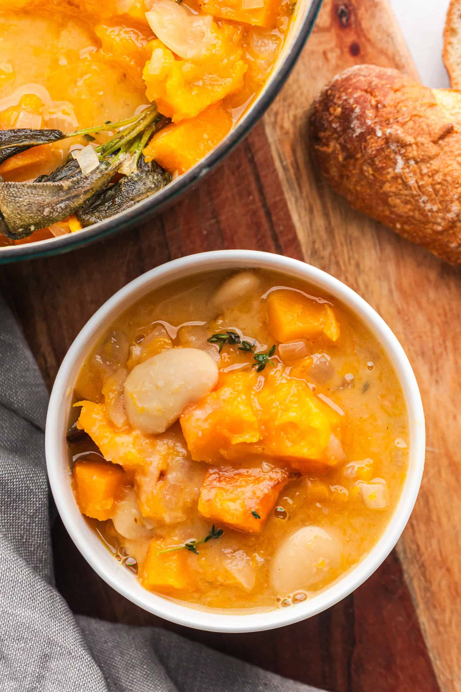 A bowl of chunky soup with white beans and orange veggies, topped with thyme. A pot and crusty bread sit behind it, showing a warm, homemade meal on a wooden table.