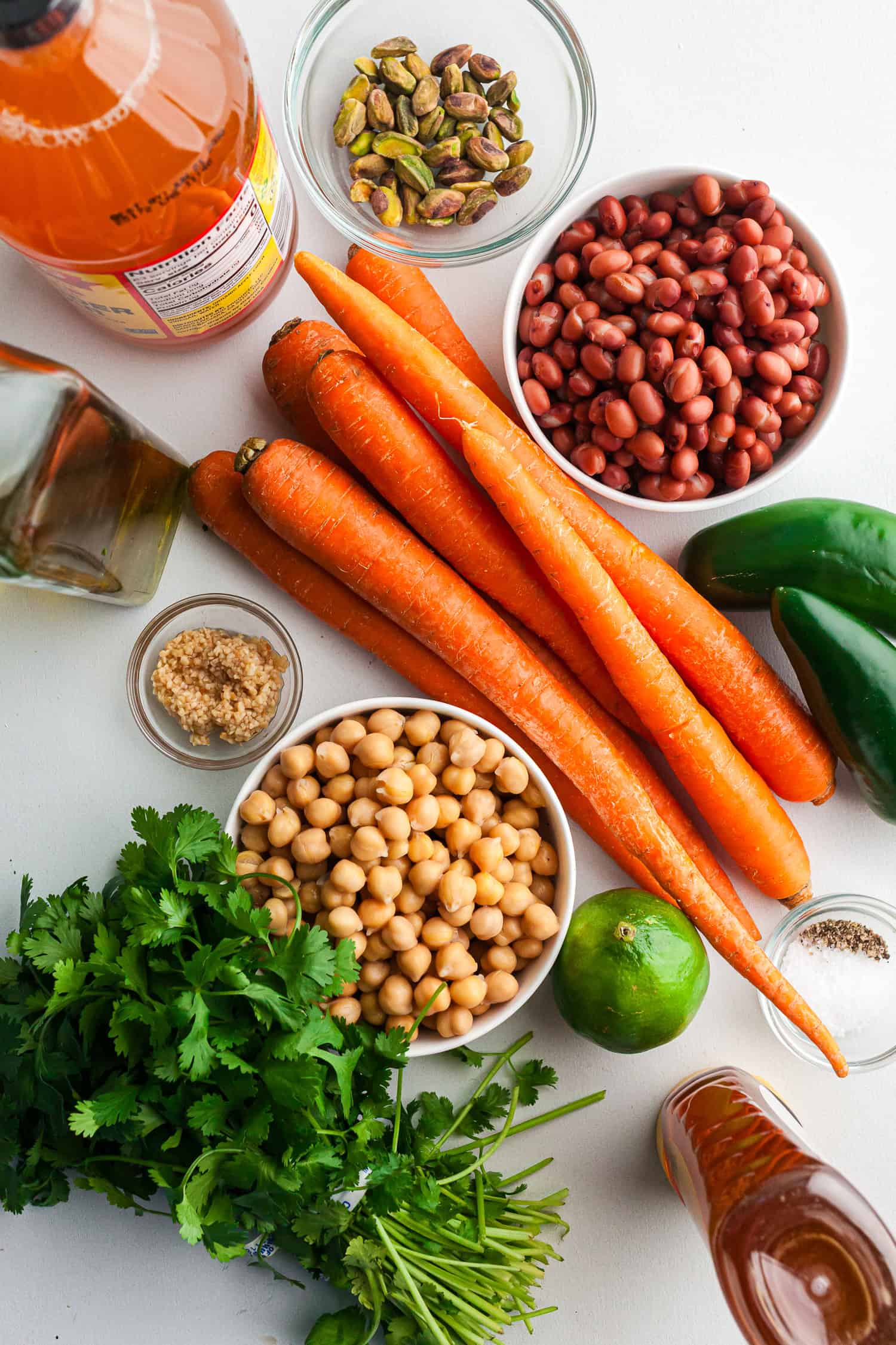 Ingredients for a fresh salad arranged on a white surface, including carrots, chickpeas, red beans, pistachios, jalapeño, lime, cilantro, garlic, olive oil, vinegar, and seasonings.