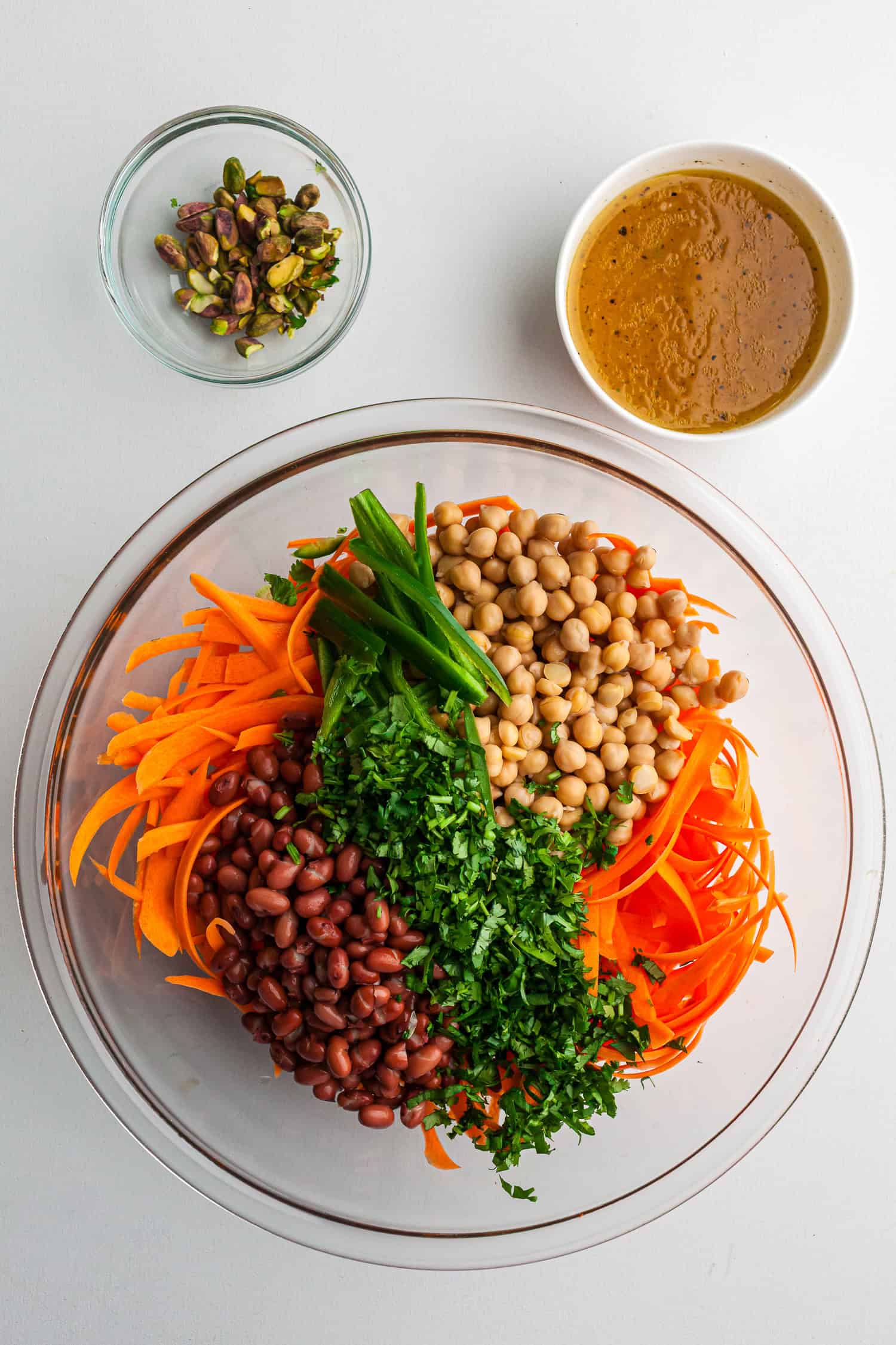 Top-down view of a glass bowl filled with shredded carrots, chickpeas, kidney beans, green onions, and chopped cilantro. Small bowls of pistachios and salad dressing are placed nearby on a white surface.