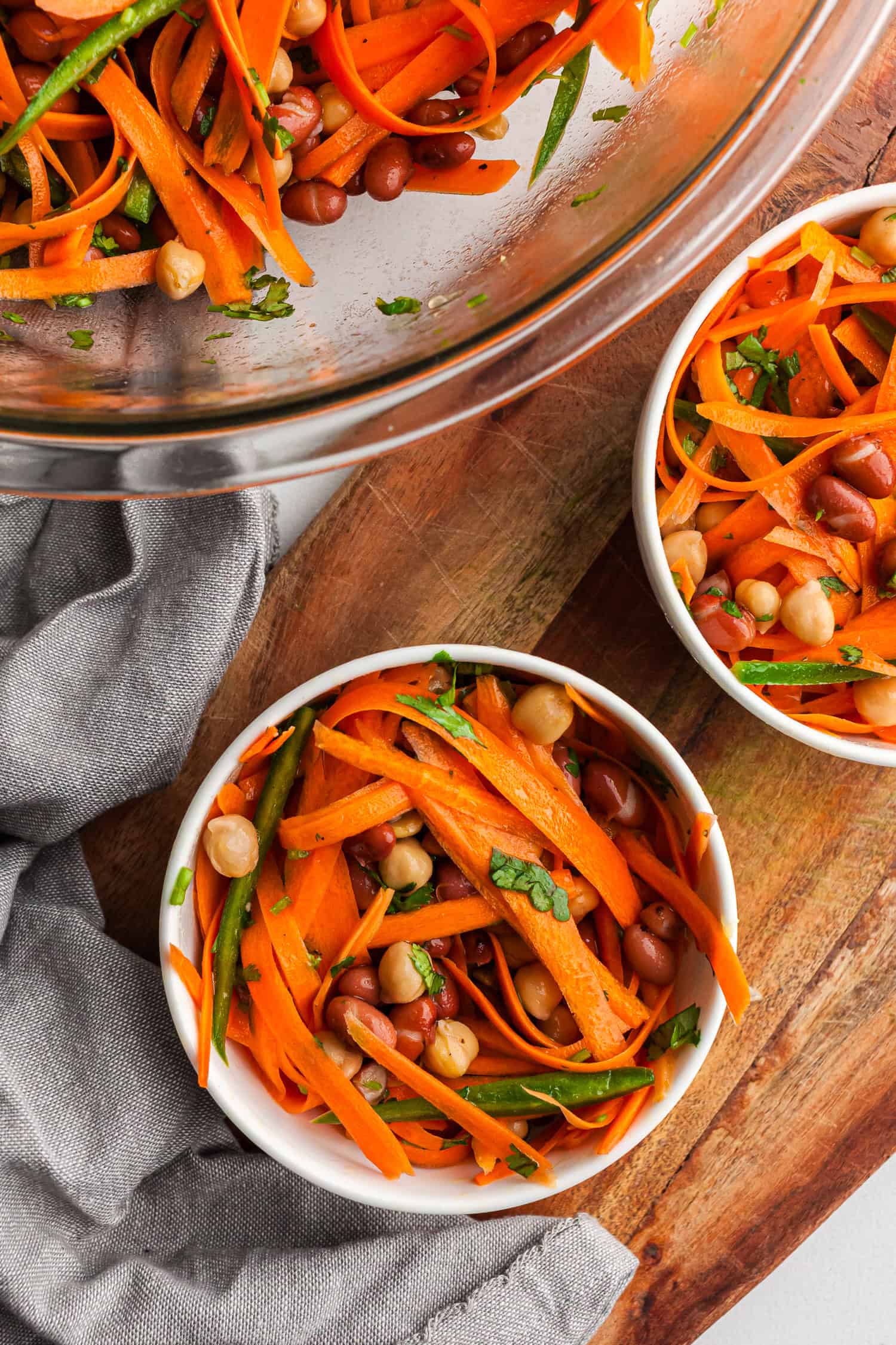 Small white bowls filled with fresh salad made of julienned carrots, chickpeas, kidney beans, and chopped cilantro. A larger glass bowl with more salad is in the background.