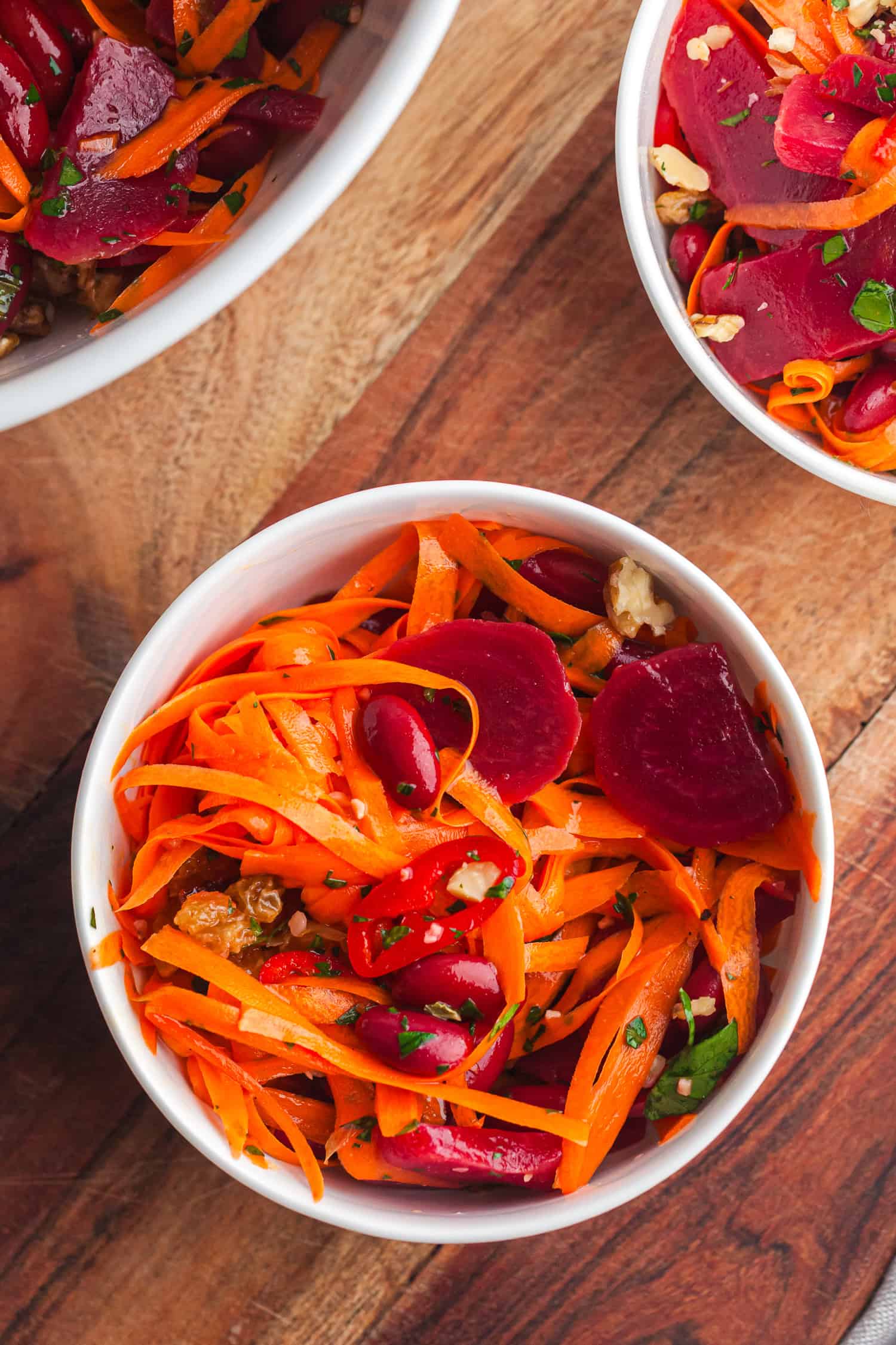 Carrot beet salad in white bowls with sliced carrots, beets, kidney beans, parsley, and walnuts on wooden table – healthy vegan meal