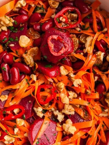 Colorful carrot beet salad with shredded carrots, kidney beans, beetroot, red chili slices, walnuts, golden raisins, and fresh herbs in a white bowl – healthy vegan recipe