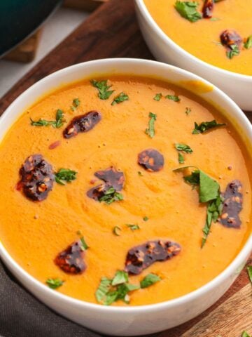 Two bowls of smooth orange carrot lentil soup with fresh green herbs and spicy chili oil on top, served on a wooden table.