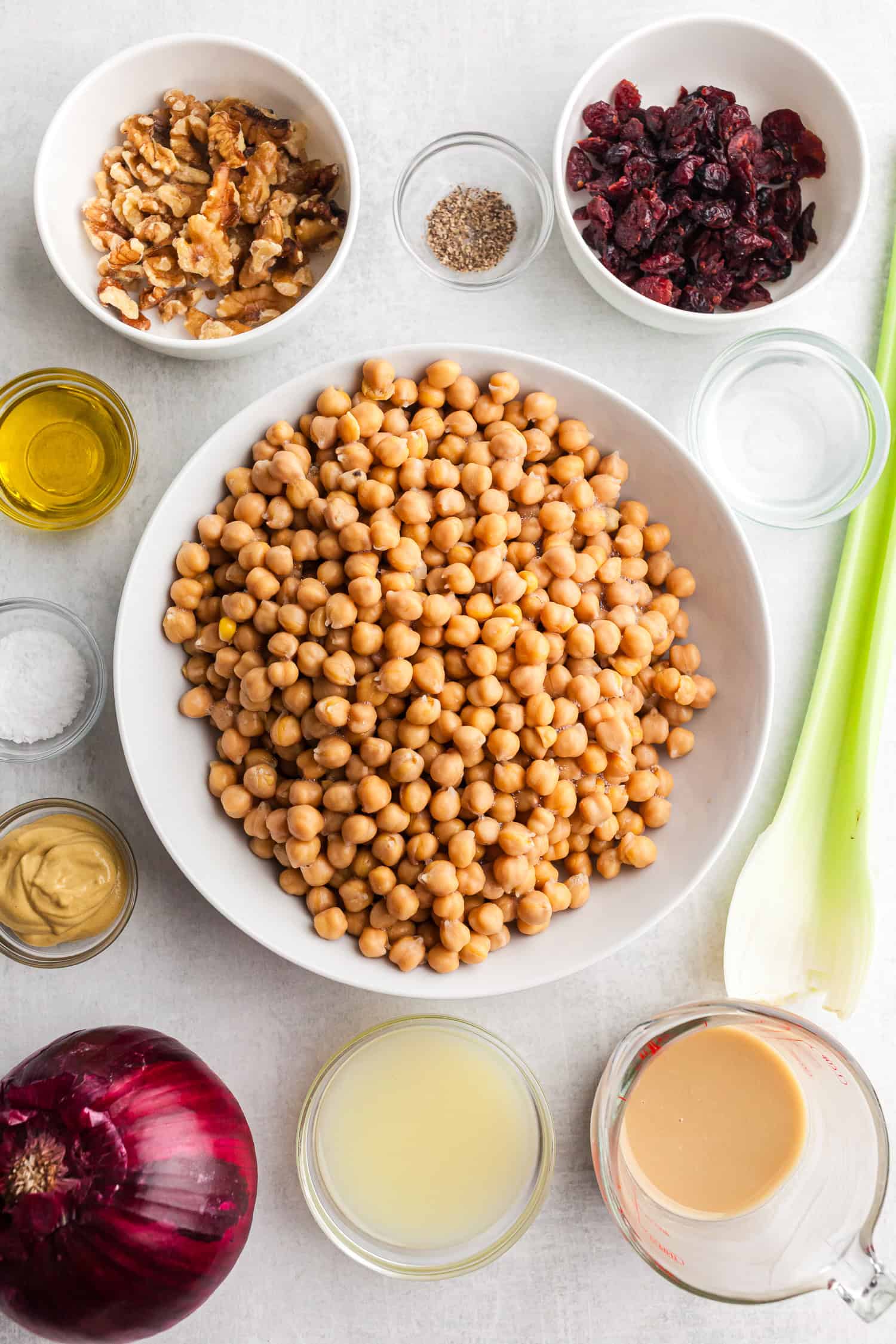 Chickpea chicken salad ingredients on white table—chickpeas, walnuts, cranberries, onion, celery, and dressing items.