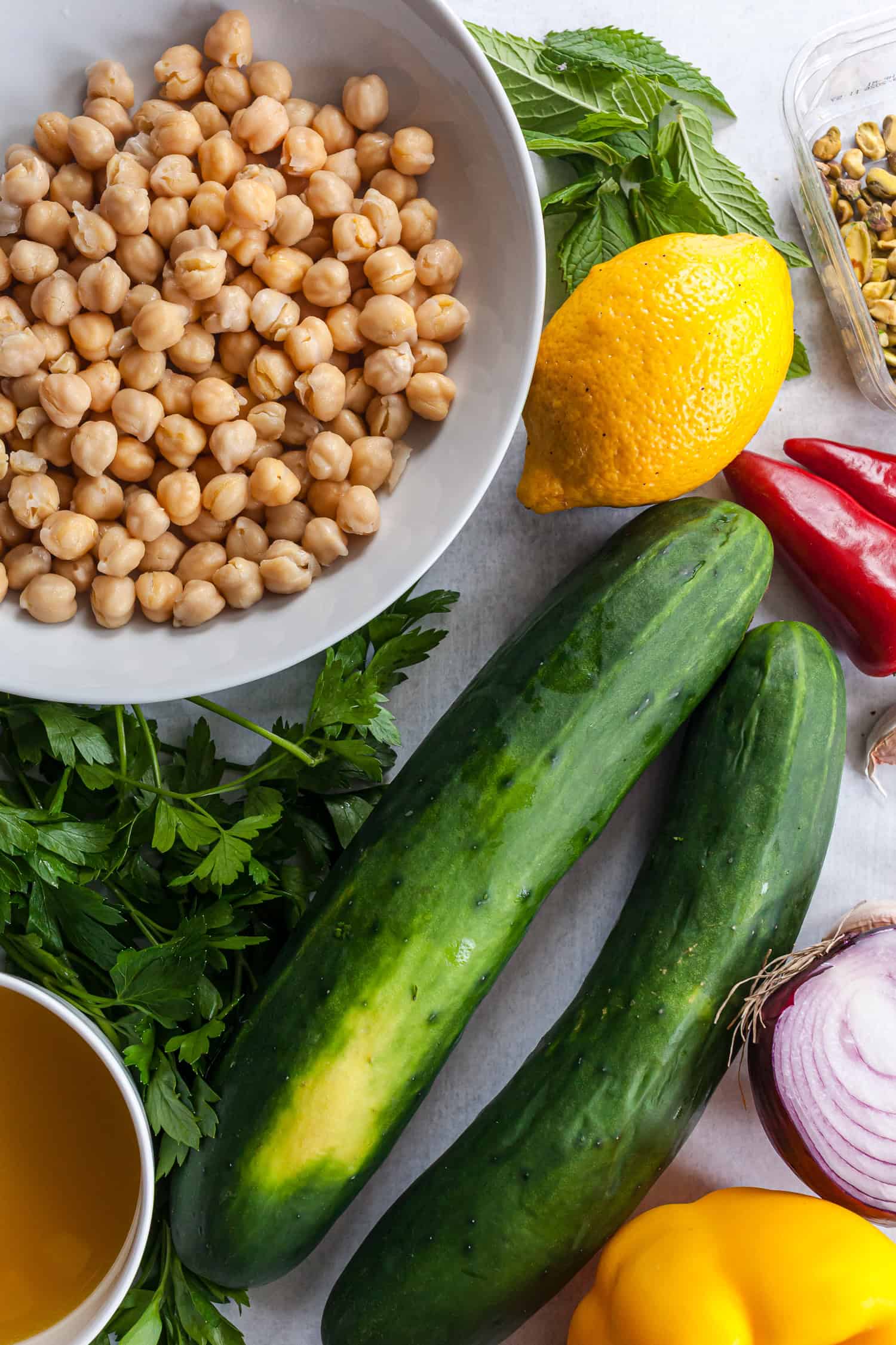 Fresh ingredients for cucumber and chickpea salad on a light surface, including chickpeas, cucumbers, lemon, mint, parsley, pistachios, chili, bell pepper, onion, and olive oil.