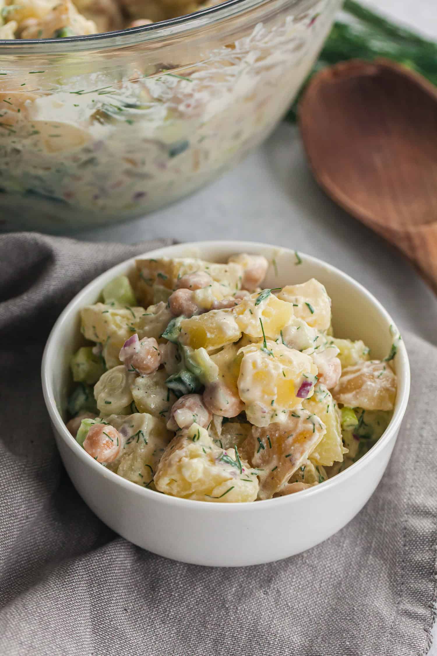 Bowl of creamy Dill Potato Salad with cooked potato chunks, chickpeas, chopped celery, red onions, and fresh dill, coated in a creamy dressing, placed on a gray cloth napkin with a larger glass bowl.