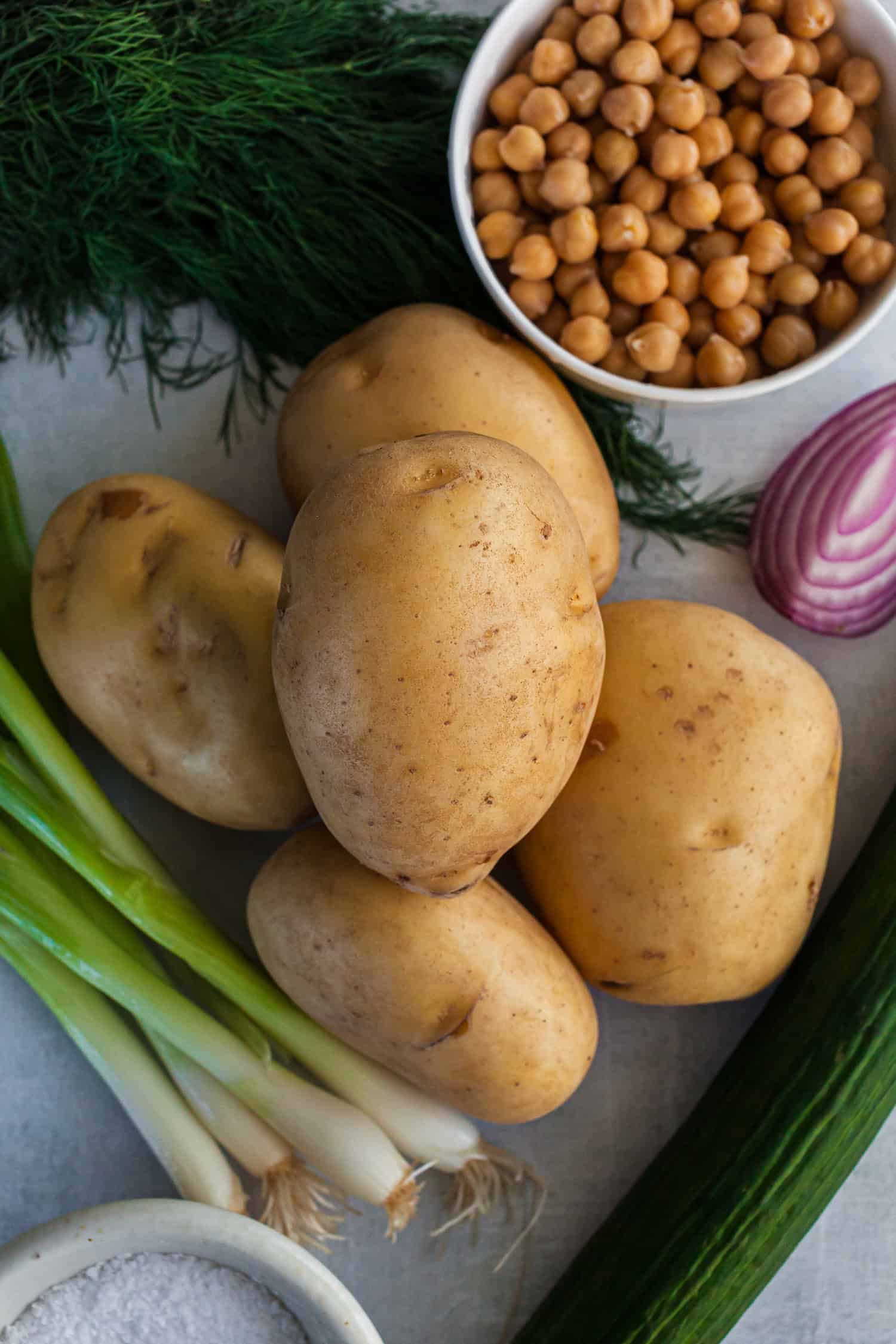 Fresh ingredients for Dill Potato Salad arranged on a light surface, including whole yellow potatoes, a bowl of chickpeas, fresh dill, halved red onion, green onions, a cucumber, and a small dish of coarse salt.