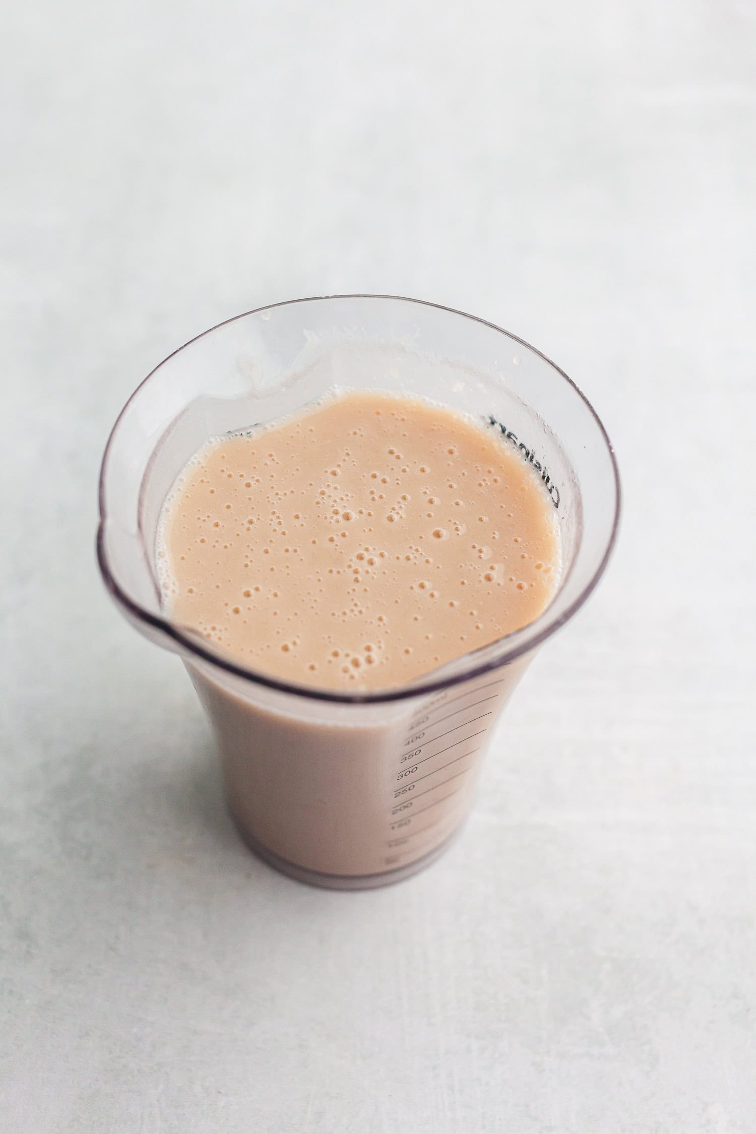 Measuring cup with blended peanut mixture, smooth and light brown, on a pale surface.