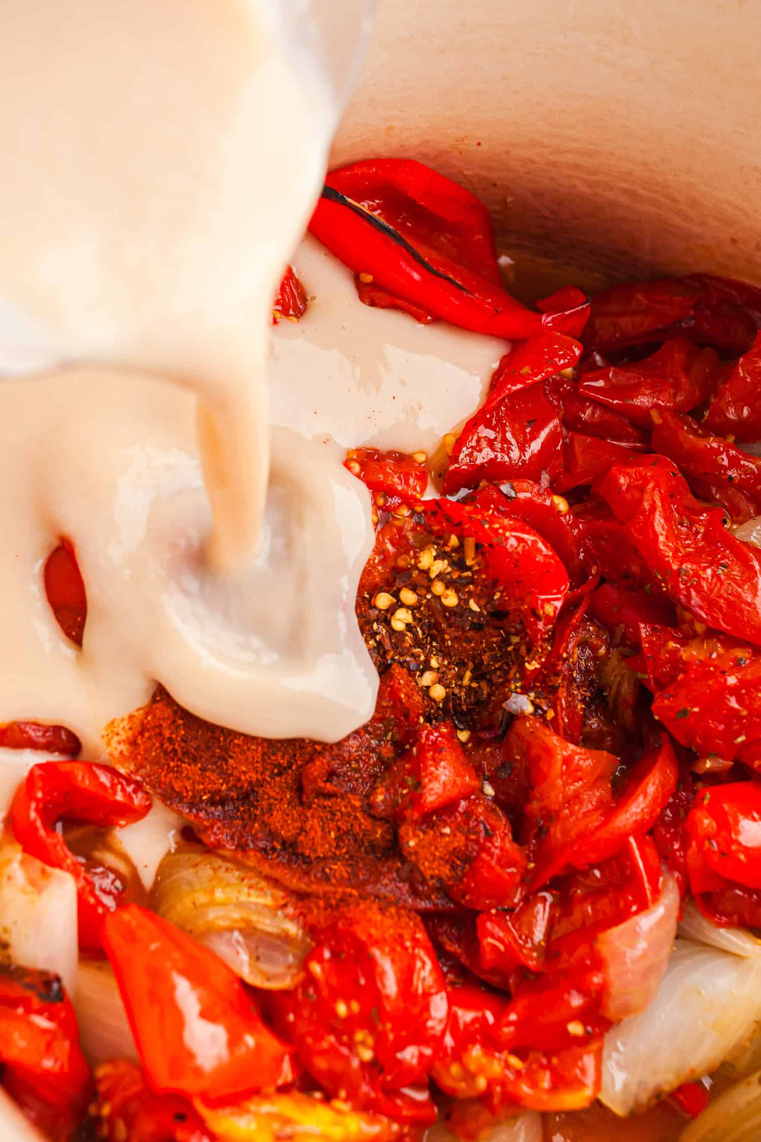 Roasted peppers, tomatoes, onions, and spices in a pot with creamy sauce being poured in.