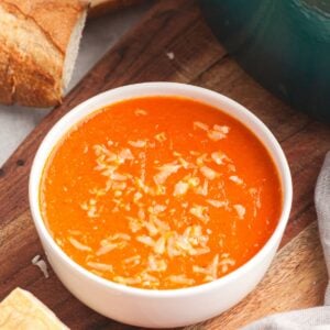 Bowl of roasted pepper and tomato soup topped with cheese, served with crusty bread on a wooden table.