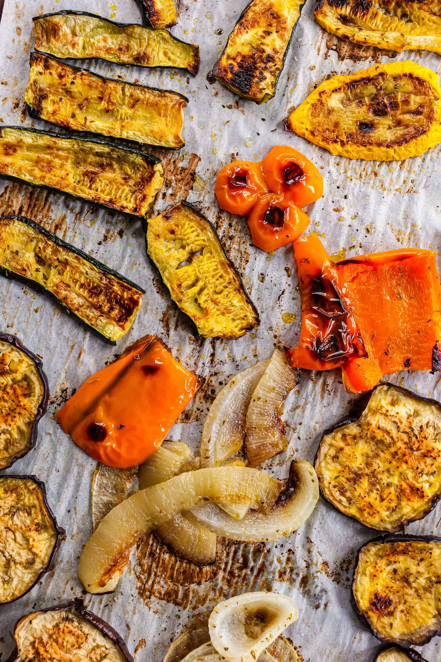 Roasted slices of zucchini, yellow squash, eggplant, red and orange bell peppers, and onion arranged on a parchment-lined baking sheet. The vegetables are evenly browned and charred, showcasing a colorful, healthy mix for a plant-based sandwich filling.