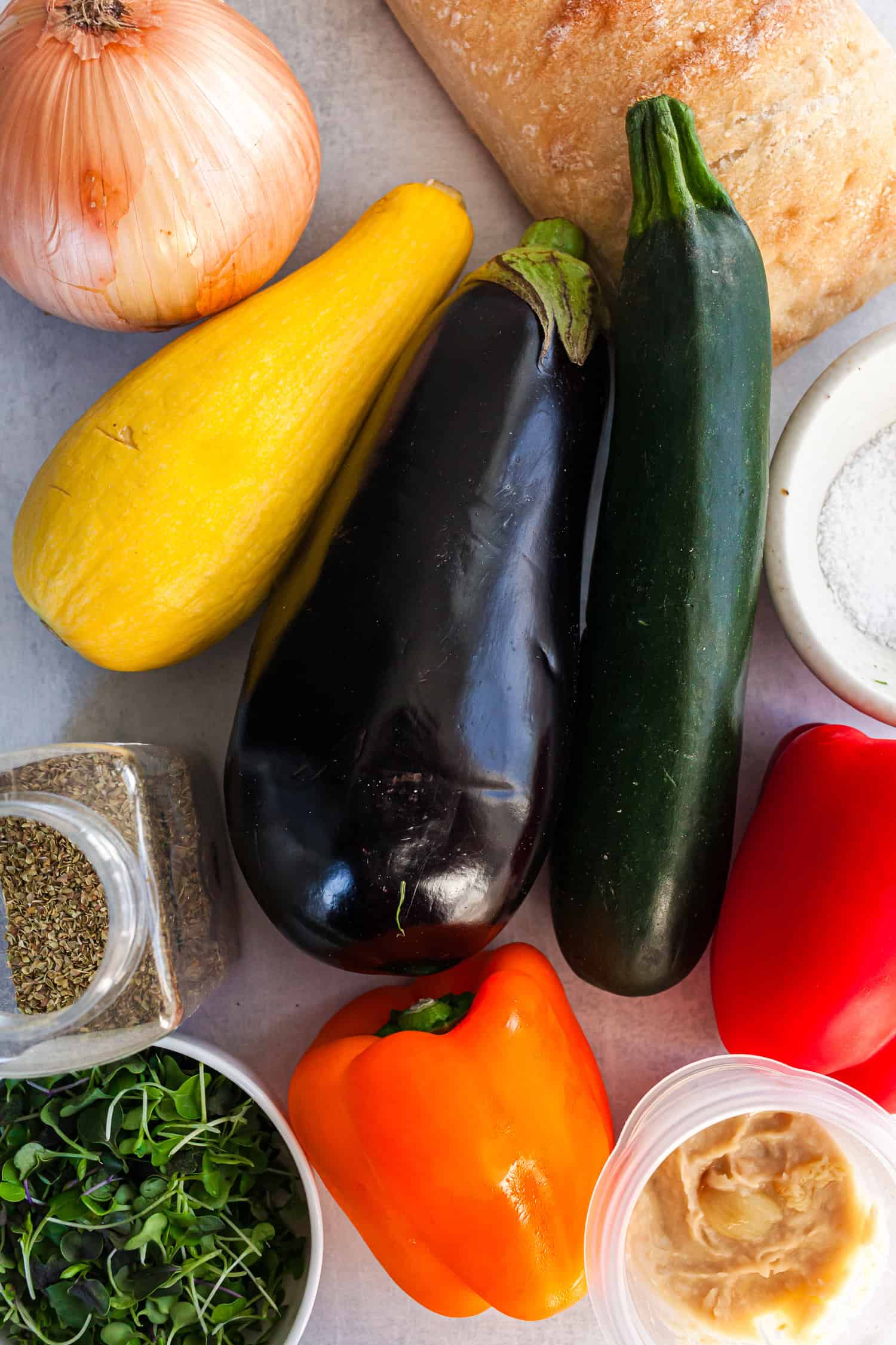 Assorted fresh ingredients for a roasted veggie sandwich displayed on a light surface, including zucchini, eggplant, yellow squash, red and orange bell peppers, onion, ciabatta bread, microgreens, hummus, salt, and dried oregano.