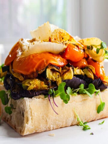 Close-up of a roasted veggie sandwich on rustic ciabatta bread, layered with grilled zucchini, roasted eggplant, red bell peppers, fresh microgreens, and cheese slices. The vegetables are tender and colorful, making the sandwich look fresh and appetizing.