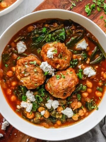 A white bowl filled with a vibrant tomato-based stew containing three large turkey meatballs, chickpeas, and wilted spinach.