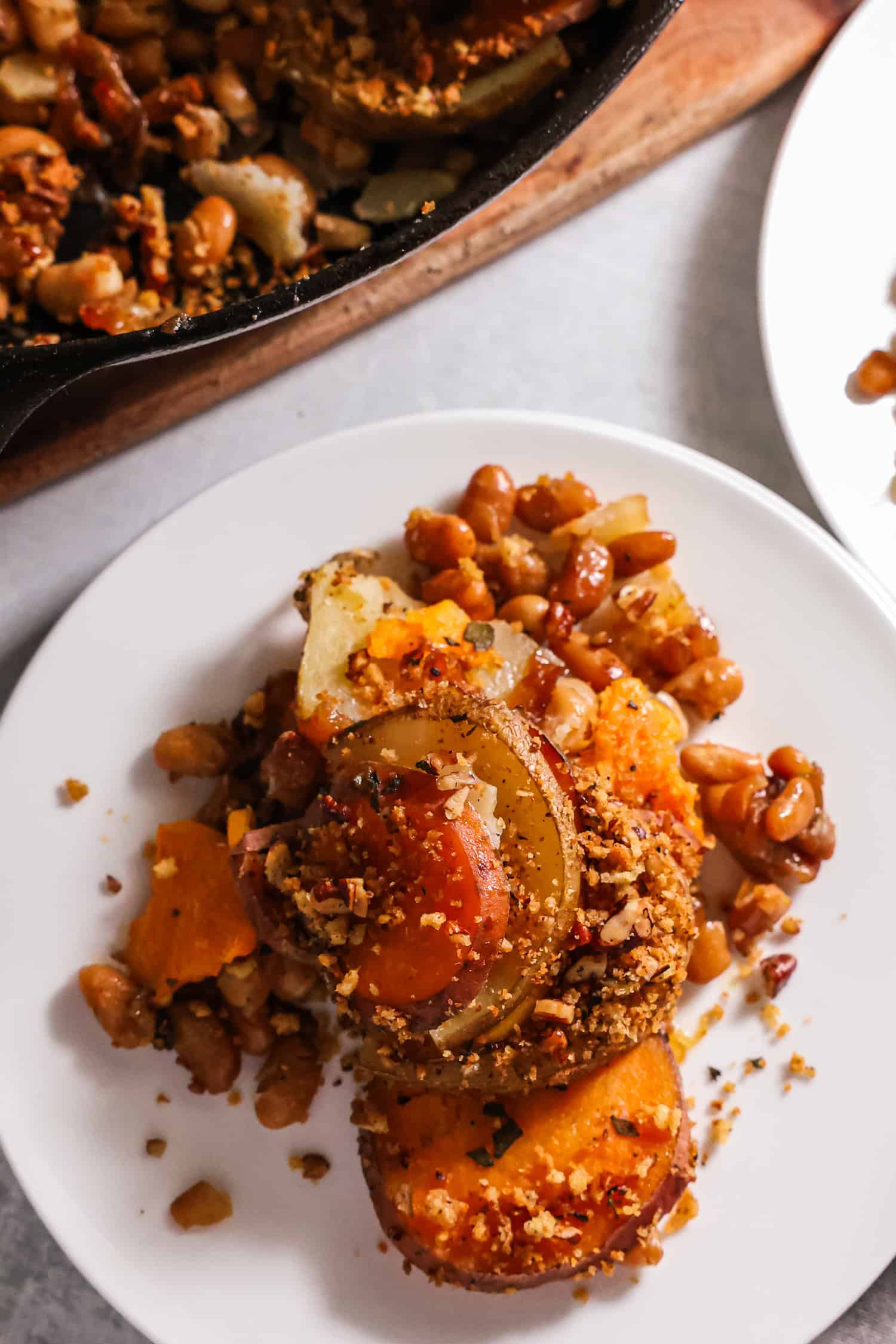 A top-down view of a cast-iron skillet filled with a spiral of sliced sweet potatoes and russet potatoes topped with a golden pecan-panko crust and fresh herb sprigs.