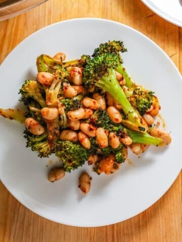 A white plate topped with roasted broccoli florets featuring charred edges and creamy white beans tossed in a vibrant red chili oil.