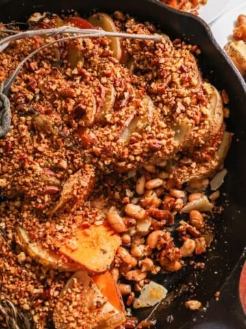 A rustic cast-iron skillet filled with a spiral of sliced russet and sweet potatoes topped with a golden-brown pecan-panko crust and nestled over a bed of white beans and fresh herbs.