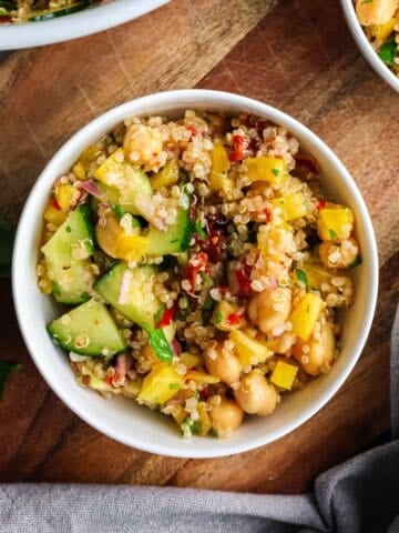 A top-down view of a white bowl filled with quinoa garbanzo bean salad, featuring chickpeas, diced cucumbers, yellow bell peppers, and fresh herbs on a wooden cutting board.
