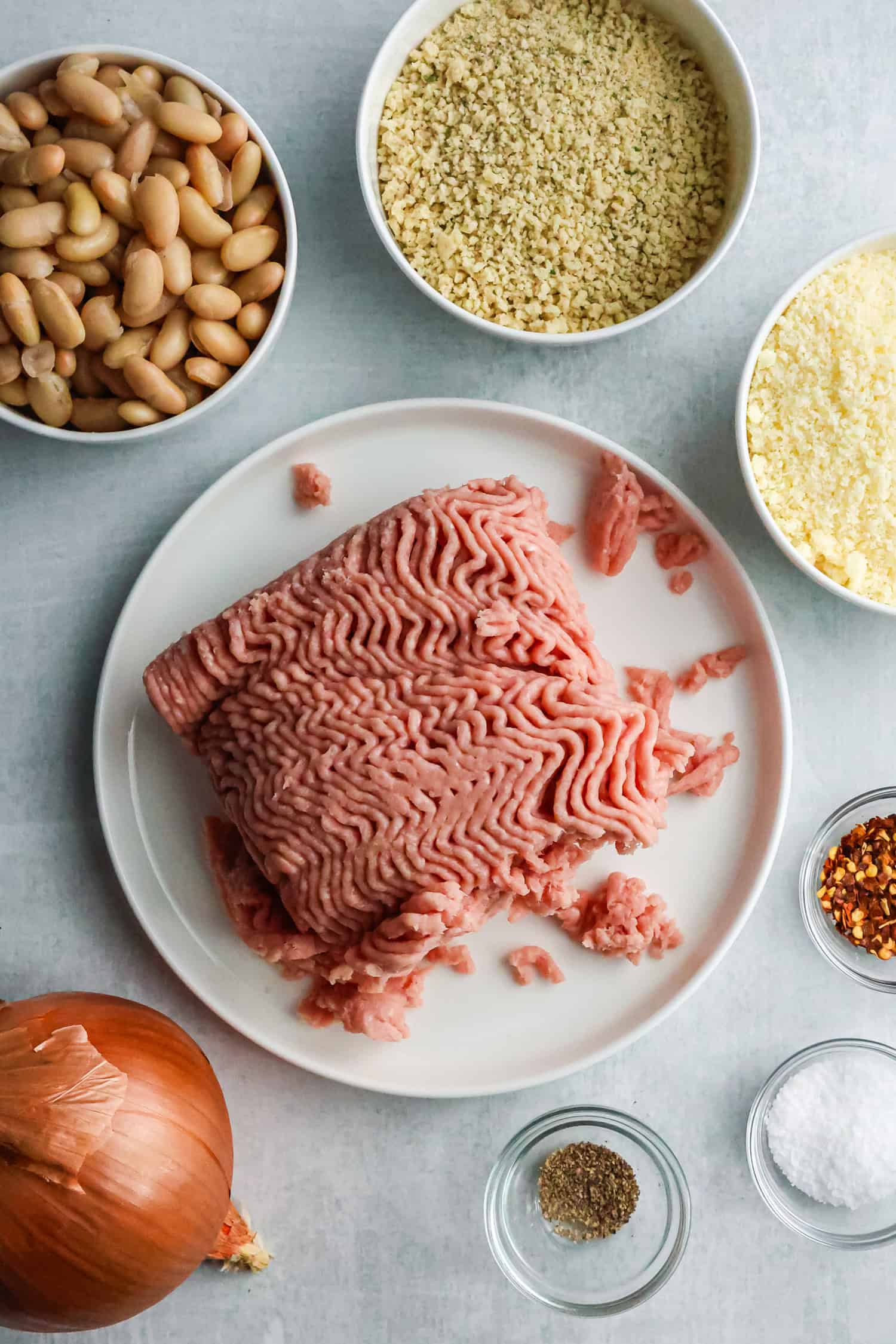 An overhead shot of fresh ingredients for chicken meatballs including ground chicken, white beans, breadcrumbs, parmesan cheese, and various spices.