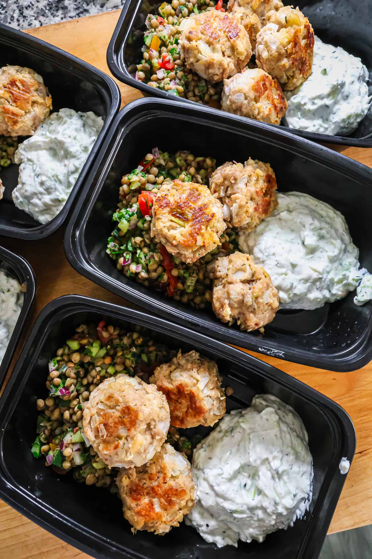 Four black meal prep containers filled with golden chicken meatballs, a fresh lentil salad, and a side of creamy white sauce.