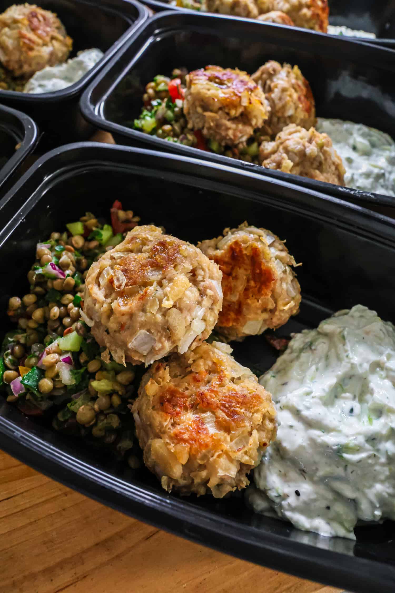 A close-up view of golden chicken meatballs, lentil salad, and a creamy dip packed inside black meal prep containers.