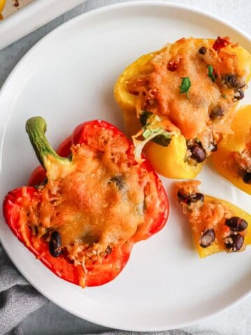 A top-down view of a red and a yellow bell pepper halved and stuffed with a mixture of black beans, quinoa, and melted golden-brown cheese on a white plate.