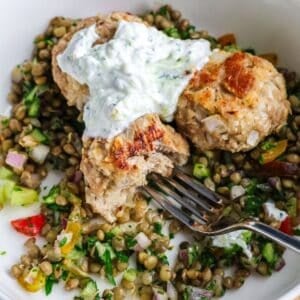 A close-up of golden-brown chicken meatballs served over a colorful lentil salad and topped with a creamy white yogurt sauce.