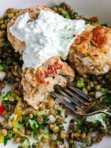 A close-up of golden-brown chicken meatballs served over a colorful lentil salad and topped with a creamy white yogurt sauce.