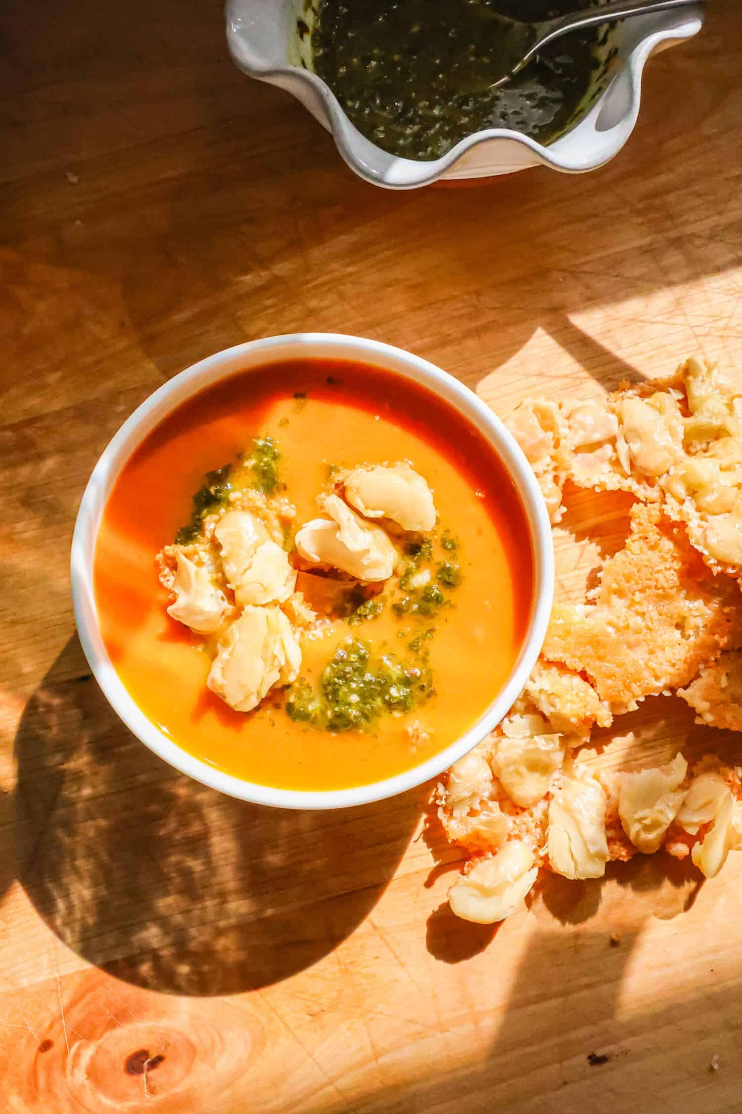 A top-down view of a white bowl filled with creamy orange tomato soup topped with green pesto and crispy butter beans, sitting next to more crunchy bean clusters on a wooden surface.