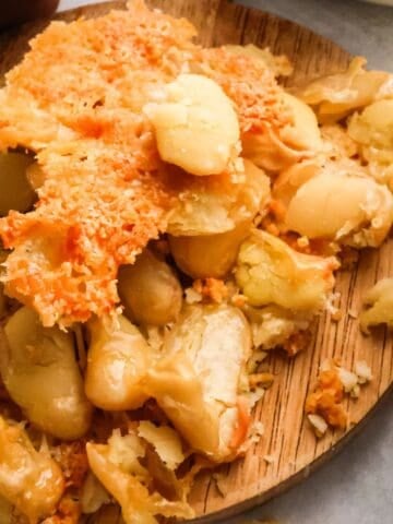 A top-down view of a white bowl of orange tomato soup topped with green pesto and roasted butter beans next to crispy bean clusters on a wooden board.