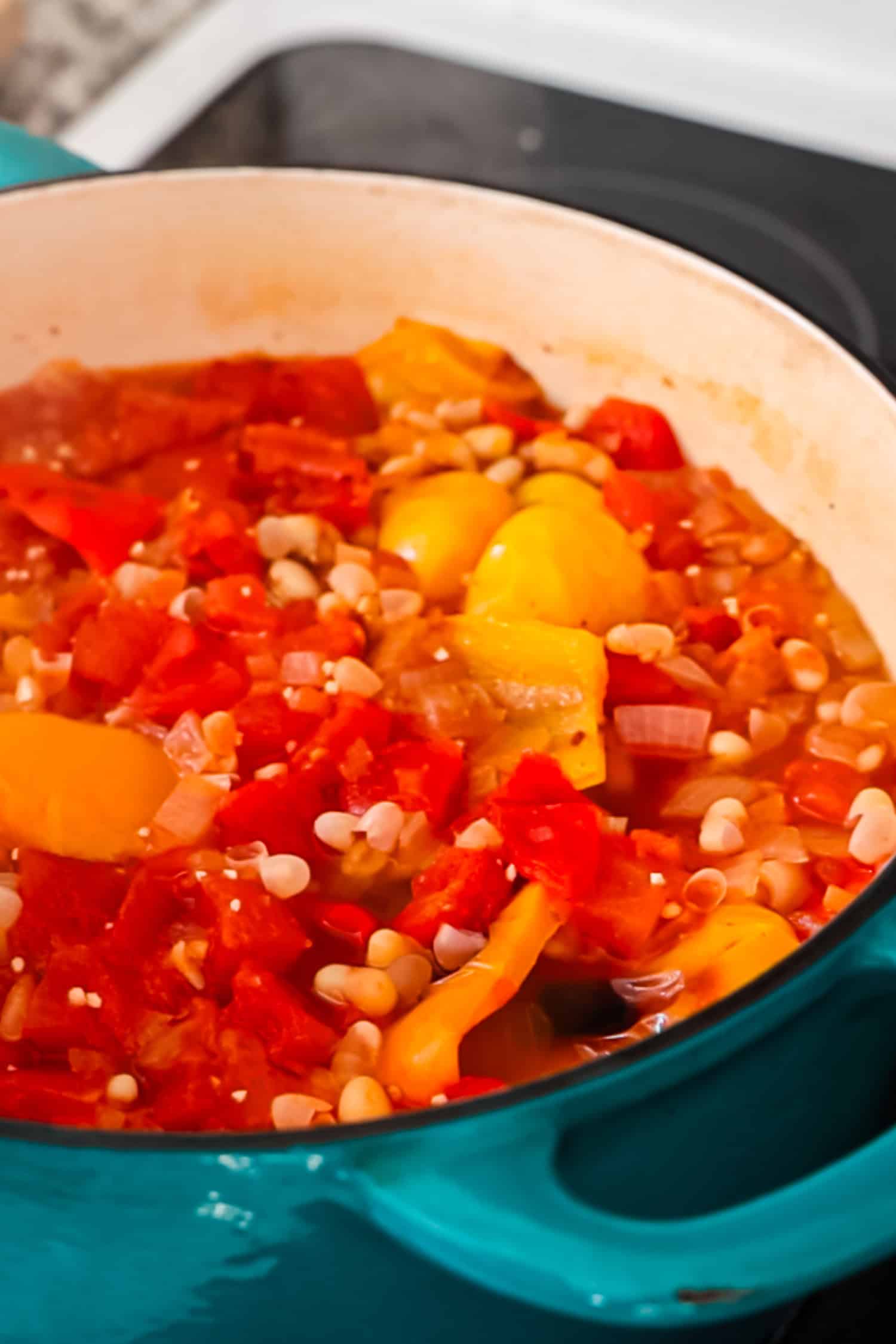 A teal Dutch oven filled with a chunky mixture of simmered tomatoes, white beans, and roasted peppers on a stovetop.